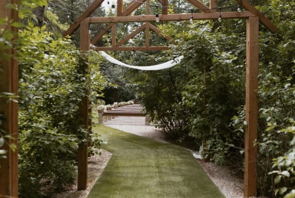 Wooden garden archway leads to outdoor wedding venue, surrounded by lush greenery and a green grass aisle.