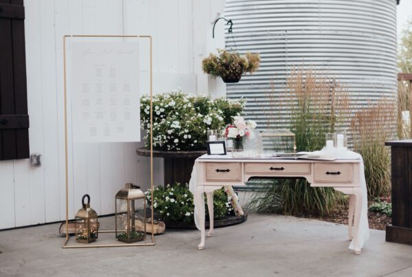 Rustic wedding decor with seating chart, lanterns, flowers, and guestbook on vintage table beside greenery backdrop.