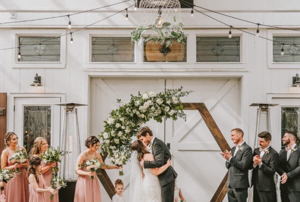 Elegant wedding ceremony with bride and groom kissing, surrounded by bridesmaids and groomsmen in a decorated venue.