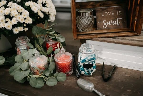 Assorted candies in glass jars with flowers and Love is sweet sign at rustic wedding candy table.