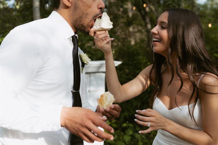 Bride and groom laughing while feeding each other cake at an outdoor wedding.